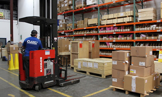 A warehouse worker in a blue Clancy shirt operating a red stand-up forklift