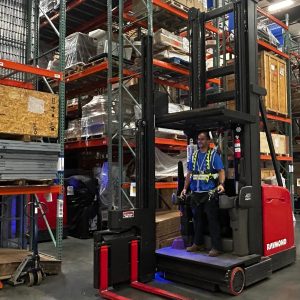 a worker wearing a safety harness operates a red Raymond forklift