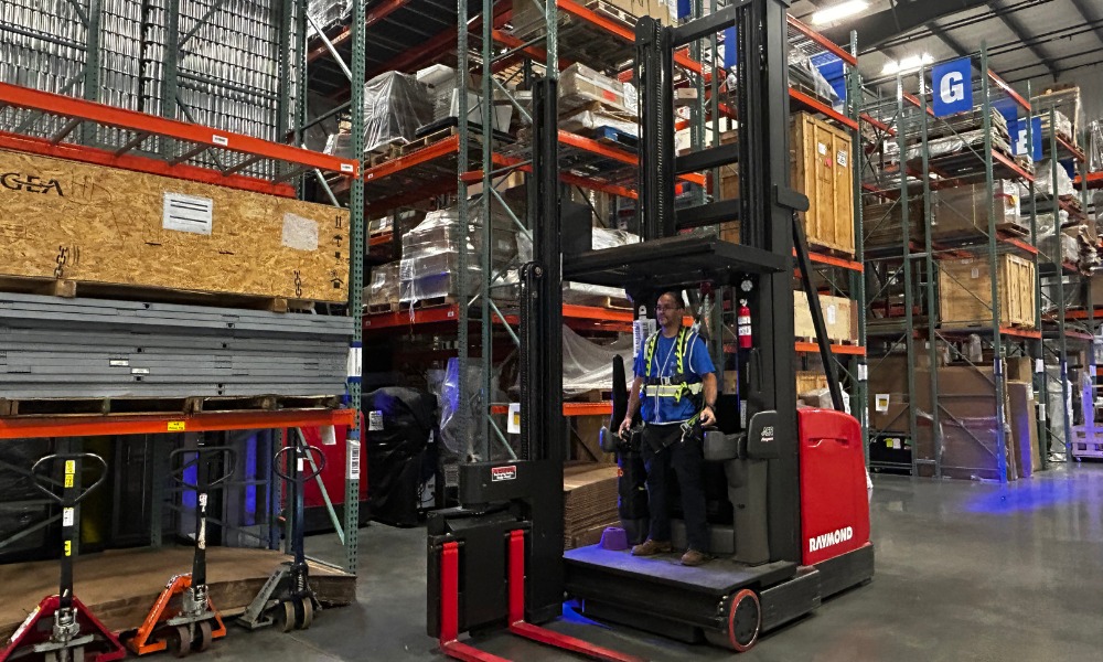 a worker wearing a safety harness operates a red Raymond forklift